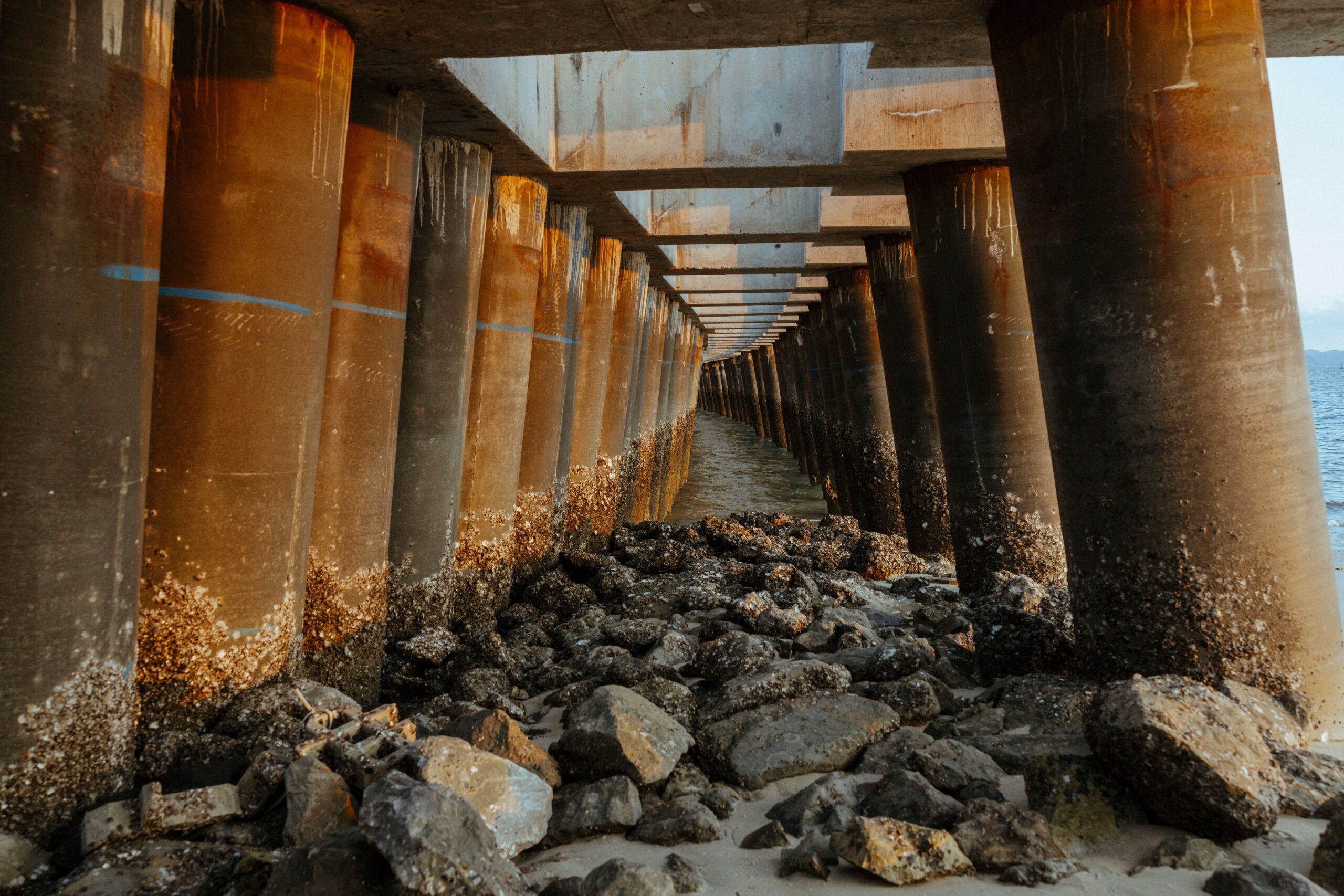 Underneath view of a seaside pier with rusted pillars and rocky shoreline, showcasing architectural symmetry.